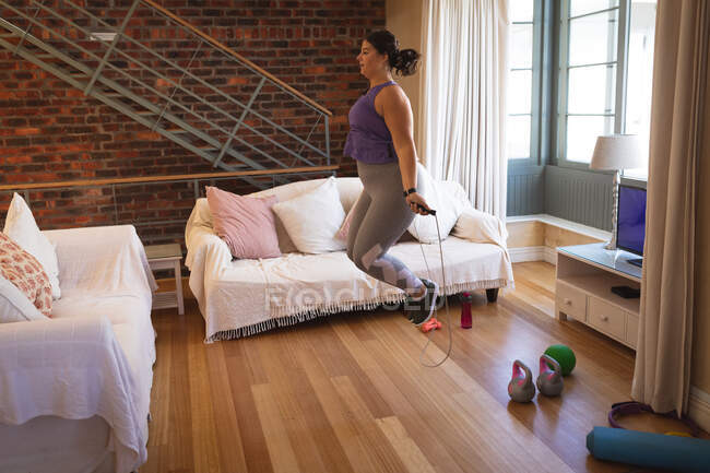 Femme vlogger caucasienne à la maison dans son salon, démontrant des exercices avec une corde à sauter pour son blog en ligne. Distance sociale et isolement personnel en quarantaine. — Photo de stock