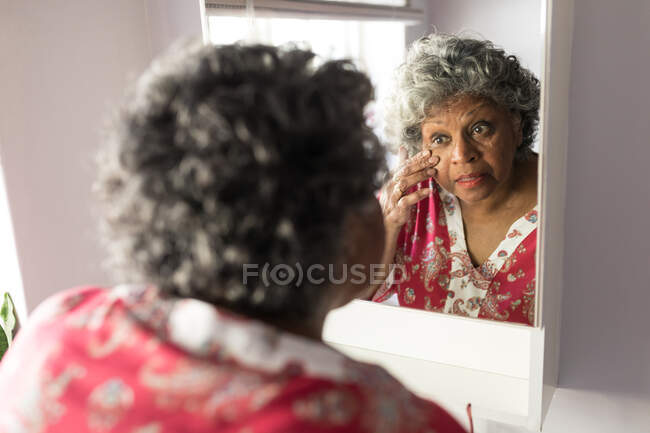 Una donna afroamericana anziana che trascorre del tempo a casa, distanziandosi socialmente e isolandosi in quarantena durante l'epidemia di coronavirus covid 19, guardandosi allo specchio e toccandosi il viso — Foto stock