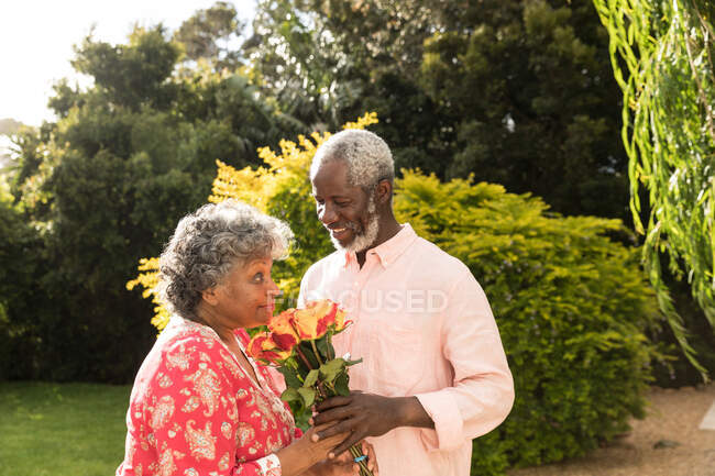 Старшая афроамериканская пара проводит время в своем саду вместе, социальное дистанцирование и самоизоляция в карантинной изоляции во время эпидемии коронавируса, мужчина, держащий букет цветов, улыбающийся и дающий их женщине — стоковое фото
