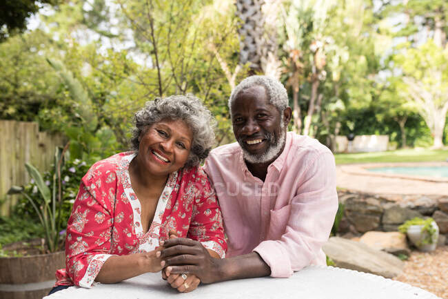 Портрет пожилой афроамериканской пары, проводящей время в своем саду вместе, социальной дистанцированности и самоизоляции в карантинной изоляции во время эпидемии коронавируса ковида 19, держащейся за руки, смотрящей в камеру и улыбающейся — стоковое фото