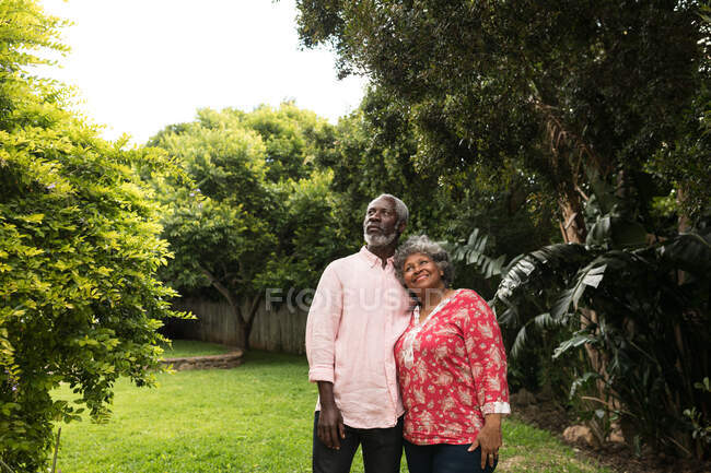 Старшая афро-американская пара проводит время в своем саду вместе, социальное дистанцирование и самоизоляция в карантинной изоляции во время эпидемии коронавируса ковируса 19, обнимаясь и отводя взгляд — стоковое фото