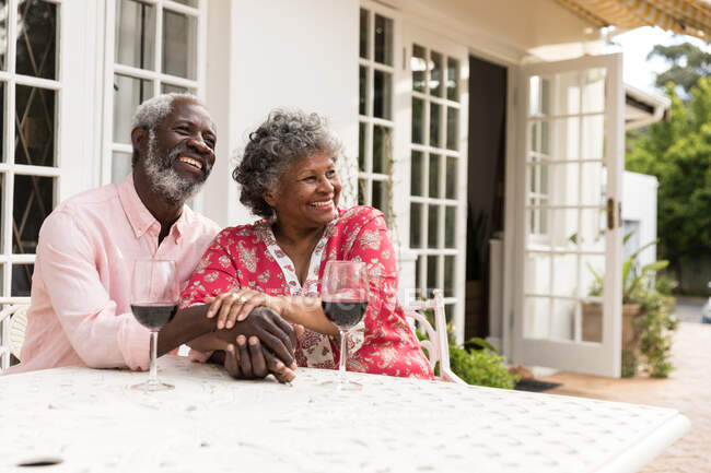 Ein älteres afroamerikanisches Paar, das während der Coronavirus-Epidemie 19 Zeit in seinem Garten verbringt, soziale Distanzierung und Selbstisolierung in Quarantäne, lächelt und schaut weg, mit einem Glas Rotwein auf dem Tisch. — Stockfoto