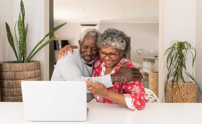 Um casal afro-americano sênior passando tempo em casa juntos, distanciamento social e auto-isolamento em quarentena durante coronavírus covid 19 epidemia, sentado a uma mesa, usando um laptop, abraçando e sorrindo — Fotografia de Stock