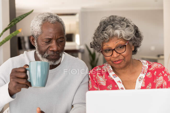 Una pareja afroamericana mayor pasa tiempo en casa juntos, distanciamiento social y aislamiento en cuarentena durante la epidemia de coronavirus covid 19, sentado en una mesa, usando una computadora portátil, el hombre sosteniendo una taza - foto de stock