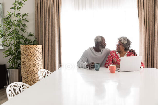 Старшая афроамериканская пара, проводящая время дома вместе, социальное дистанцирование и самоизоляция в карантинной изоляции во время эпидемии коронавируса, сидящая за столом, используя ноутбук, человек, держащий кружку — стоковое фото