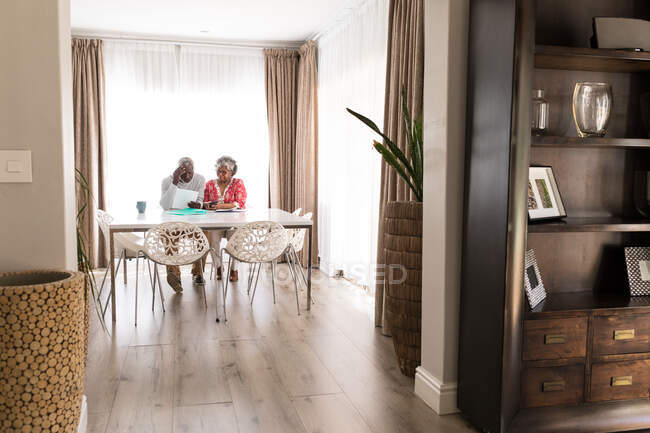 Старшая афроамериканская пара, проводящая время дома вместе, социальное дистанцирование и самоизоляция в карантинной изоляции во время эпидемии коронавируса 19-го типа, сидящая за столом, мужчина с клочком бумаги — стоковое фото