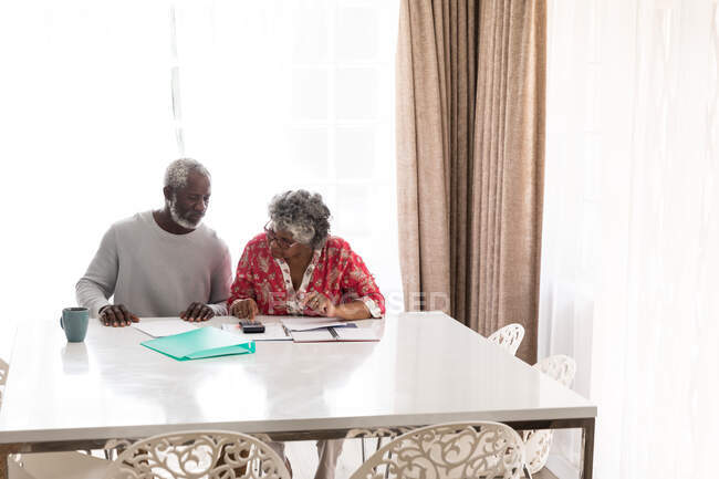 Старша афроамериканська пара проводить час удома разом, спілкуючись і самоізолюючись під час карантинної блокування під час епідемії коронавірусу, 19 років тому, сидячи за столом, використовуючи калькулятор. — стокове фото