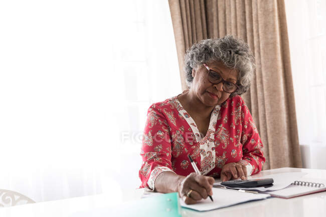 Старшая афроамериканка, проводящая время дома, социальное дистанцирование и самоизоляция в карантинной изоляции во время эпидемии коронавируса, сидя за столом, используя калькулятор и делая заметки — стоковое фото
