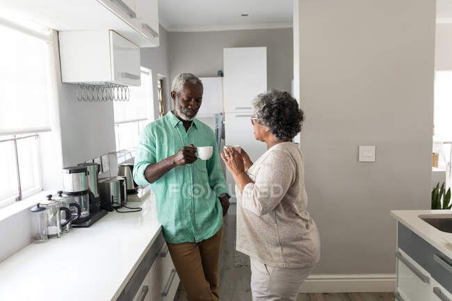 Un couple afro-américain âgé passe du temps à la maison ensemble, la distance sociale et l'isolement personnel en quarantaine pendant l'épidémie de coronavirus covid 19, debout dans la cuisine, parlant, tenant des tasses — Photo de stock