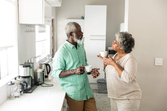 Una coppia afroamericana anziana che trascorre del tempo a casa insieme, distanziamento sociale e isolamento in quarantena durante l'epidemia di coronavirus covid 19, in piedi in cucina, parlando, tenendo le tazze — Foto stock