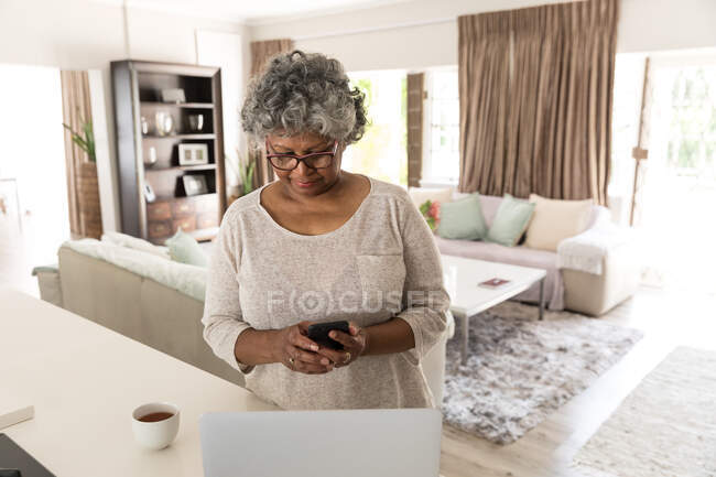 Une femme afro-américaine âgée passe du temps à la maison, à prendre des distances sociales et à s'isoler en quarantaine pendant l'épidémie de coronavirus covid 19, à l'aide d'un smartphone — Photo de stock