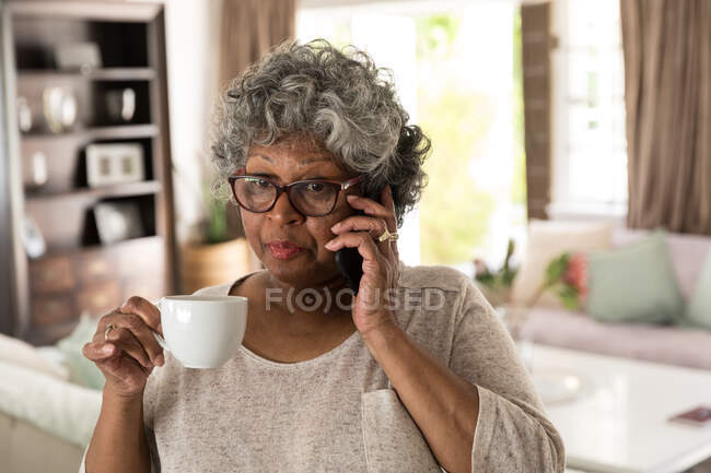 Une femme afro-américaine âgée passe du temps à la maison, à prendre des distances sociales et à s'isoler en quarantaine pendant l'épidémie de coronavirus covid 19, parlant sur un smartphone et tenant une tasse — Photo de stock