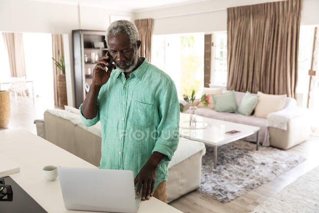 Un anziano afroamericano che trascorre del tempo a casa, allontanandosi socialmente e isolandosi in quarantena durante l'epidemia di coronavirus covid 19, parlando su uno smartphone e usando un computer portatile — Foto stock