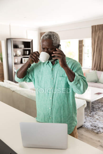 Старший афроамериканец проводит время дома, социальное дистанцирование и самоизоляция в карантинной изоляции во время эпидемии коронавируса, разговаривает по смартфону и пьет кофе. — стоковое фото