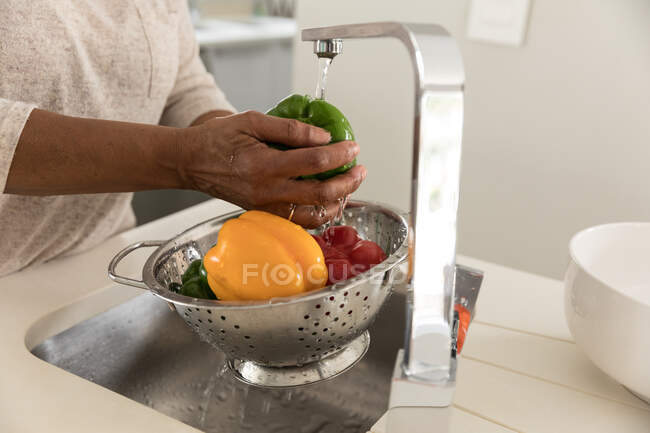 Close up mid section of African American woman spending time at home, social distancing and self isolation in quarantine lockdown during coronavirus covid 19 epidemic, holding a strainer and washing peppers — Stock Photo