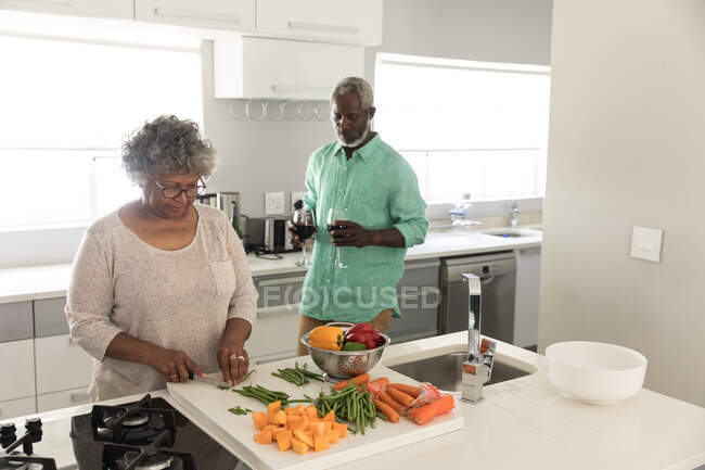 Un couple afro-américain âgé passe du temps à la maison ensemble, la distance sociale et l'isolement personnel en quarantaine pendant l'épidémie de coronavirus covid 19, la femme coupant des légumes, l'homme tenant des verres de vin rouge — Photo de stock
