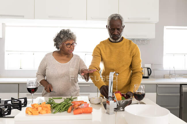 Un couple afro-américain âgé passe du temps à la maison ensemble, la distance sociale et l'isolement personnel en quarantaine pendant l'épidémie de coronavirus covid 19, l'homme qui lave les légumes, la femme qui parle — Photo de stock
