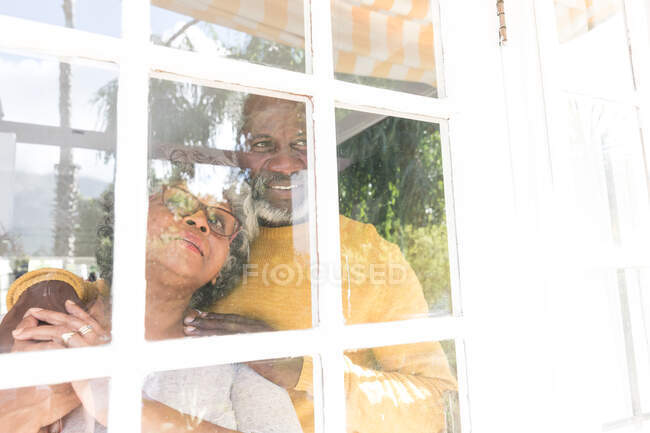 Um casal afro-americano sênior passando tempo em casa juntos, distanciamento social e auto-isolamento em quarentena durante coronavírus covid 19 epidemia, olhando para fora da janela sorrindo, abraçando — Fotografia de Stock