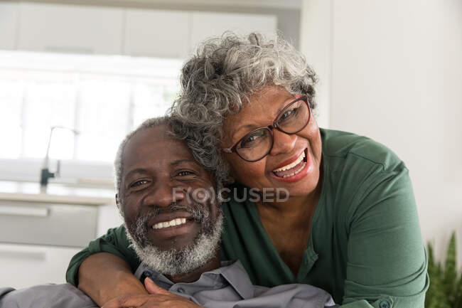Портрет високопоставленої афроамериканської пари, яка проводить час вдома разом, соціальна дистанція і самоізоляція в карантинній блокуванні під час епідемії коронавірусу (19), дивлячись на камеру, посміхаючись, обіймаючись — стокове фото