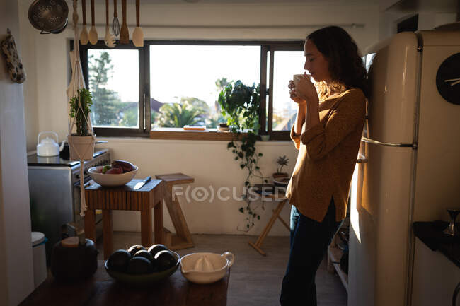 Mujer caucásica pasar tiempo en casa auto aislamiento y distanciamiento social en cuarentena de bloqueo durante coronavirus covid 19 epidemia, de pie en su cocina y tomando café. - foto de stock
