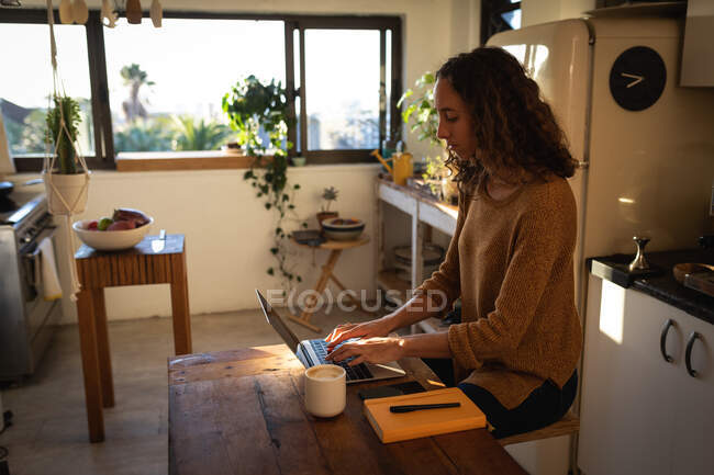 Кавказька жінка проводить вдома самоізоляцію і соціальну дистанцію в карантині під час епідемії коронавірусу covid 19, працюючи на кухні, використовуючи свій ноутбук.. — стокове фото