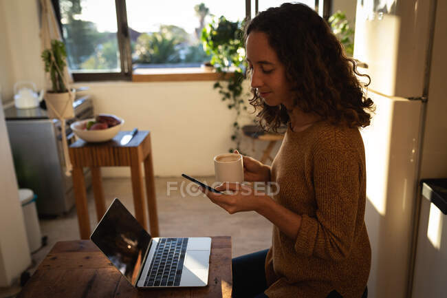 Caucasian woman spending time at home self isolating and social distancing in quarantine lockdown during coronavirus covid 19 epidemic, working in her kitchen, using her laptop computer and smartphone. — Stock Photo