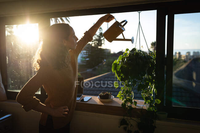 Кавказская женщина проводит время дома, изолируя себя и отдаляясь от общества в карантинной изоляции во время эпидемии коронавируса ковид 19, поливая растения на кухне. — стоковое фото