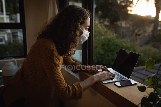 Кавказька жінка проводить час удома на самоті, одягнувши маску обличчя проти коронавірусу covid19, сидячи біля вікна і працюючи за допомогою ноутбука.. — стокове фото