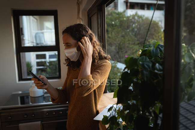 Kaukasische Frau verbringt Zeit zu Hause selbstisoliert, trägt eine Gesichtsmaske gegen Covid19 Coronavirus, steht am Fenster und arbeitet mit ihrem Smartphone. — Stockfoto