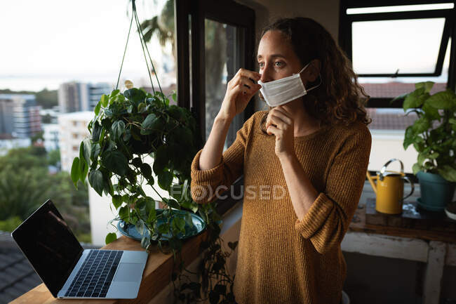 Kaukasische Frau verbringt Zeit zu Hause selbstisolierend, setzt eine Gesichtsmaske gegen Covid19 Coronavirus auf, steht am Fenster und bereitet sich mit ihrem Laptop auf die Arbeit vor. — Stockfoto