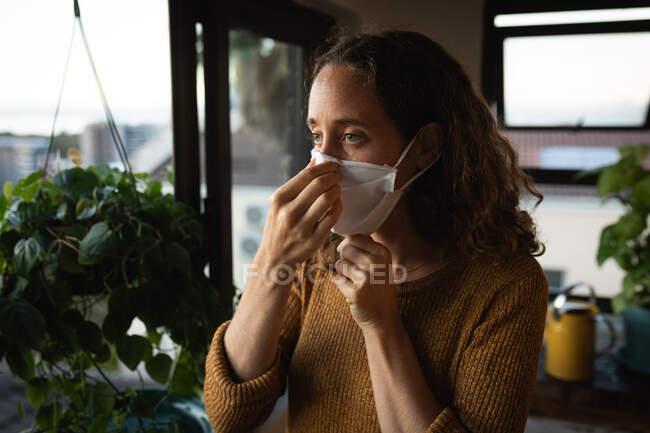 Mujer caucásica pasar tiempo en casa auto aislamiento y distanciamiento social en cuarentena bloqueo durante coronavirus covid 19 epidemia, poniendo una máscara facial contra covid19 coronavirus, de pie junto a una ventana. - foto de stock