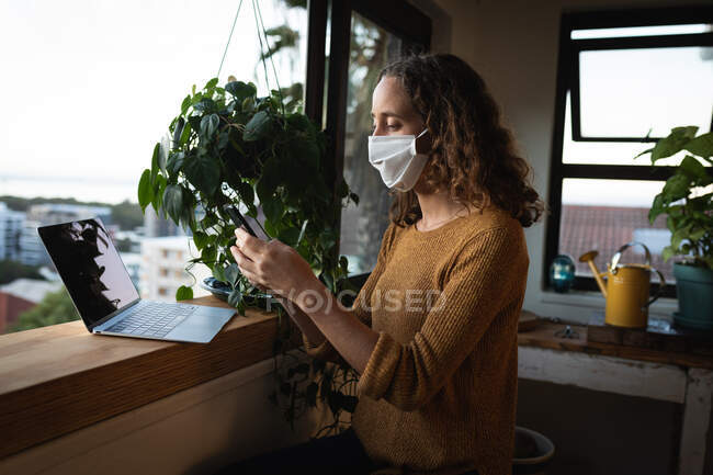 Kaukasische Frau, die ihre Zeit zu Hause selbstisolierend verbringt, eine Gesichtsmaske gegen Covid19 Coronavirus trägt, am Fenster steht, ihr Smartphone und einen Laptop benutzt. — Stockfoto