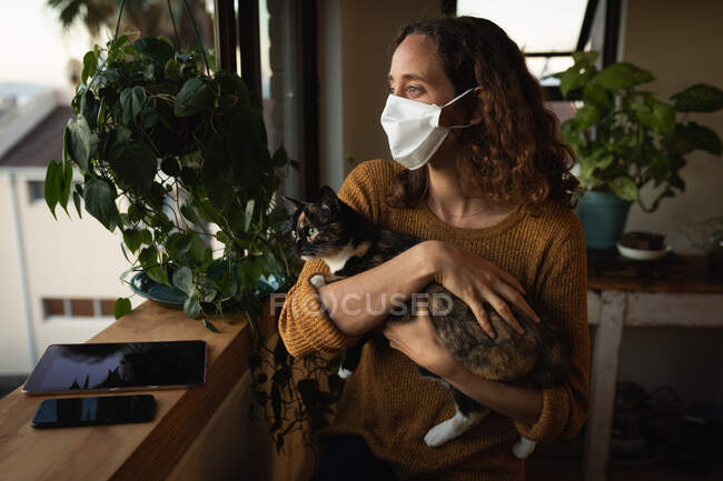 Mujer caucásica pasando tiempo en casa aislándose, usando una máscara facial contra el covid19 coronavirus, de pie junto a una ventana y sosteniendo a su gato. - foto de stock