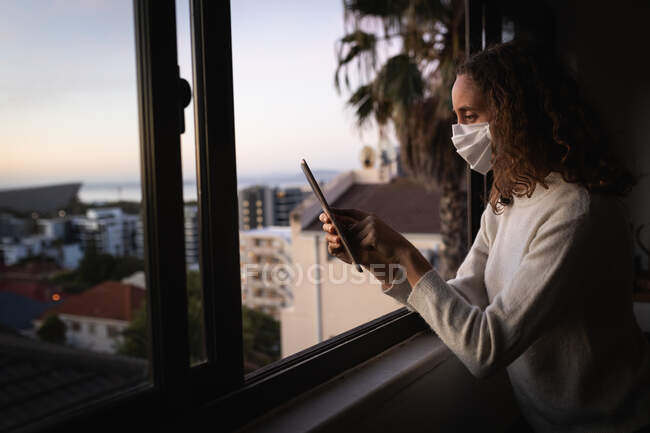 Caucasian woman spending time at home self isolating and social distancing in quarantine lockdown during coronavirus covid 19 epidemic, wearing a face mask against covid19 coronavirus, standing by a window and using her tablet. — Stock Photo