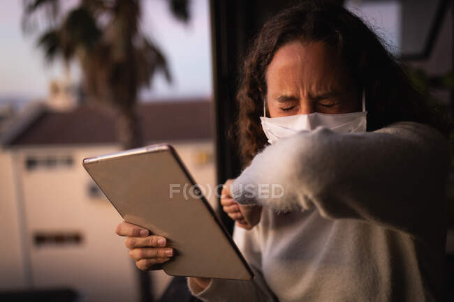 Caucasian woman spending time at home self isolating and social distancing in quarantine lockdown during coronavirus covid 19 epidemic, wearing a face mask, standing by a window, using her tablet and covering her mouth while coughing. — Stock Photo