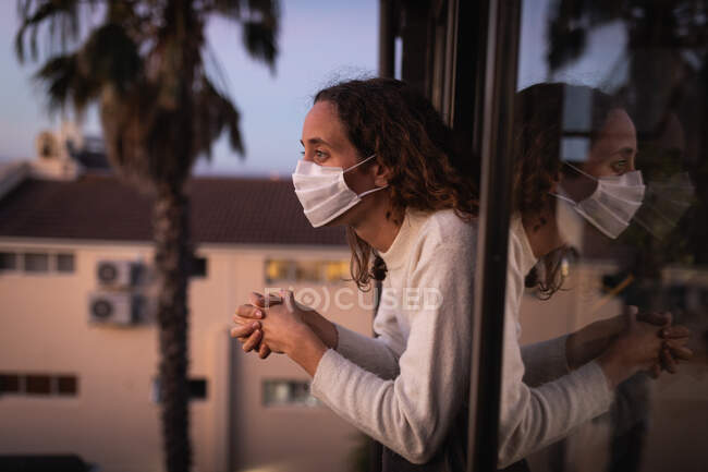 Mujer caucásica pasar tiempo en casa auto aislamiento y distanciamiento social en cuarentena de bloqueo durante coronavirus covid 19 epidemia, con una máscara facial contra covid19 coronavirus, mirando a través de la ventana. - foto de stock