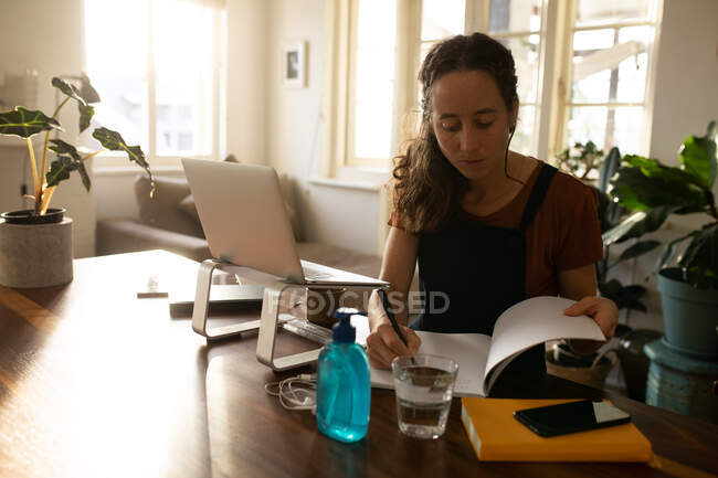 Mulher caucasiana passando tempo em casa, sentada ao lado de sua mesa e trabalhando, usando seu laptop e notebook. Distanciamento social e auto-isolamento em quarentena . — Fotografia de Stock