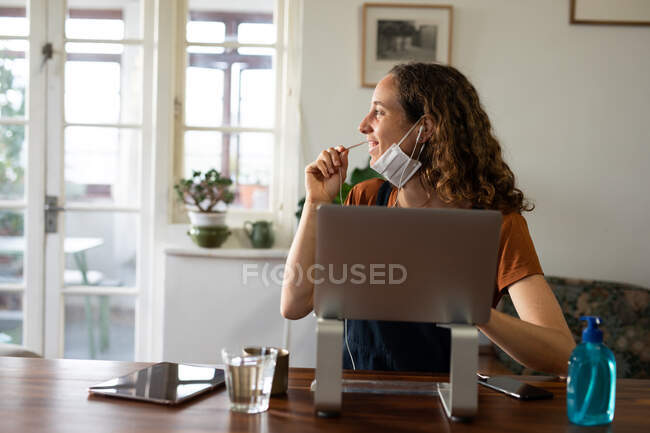 Caucasian woman spending time at home, wearing a face mask against coronavirus, covid 19, sitting by her desk and working, using her laptop and earphones. Social distancing and self isolation in quarantine lockdown. — Stock Photo