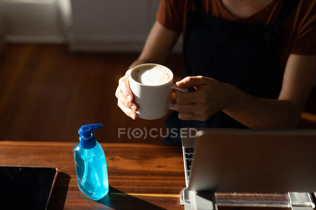 Die mittlere Hälfte der Frauen verbringt Zeit zu Hause, sitzt am Schreibtisch, macht eine Pause von der Arbeit und hält eine Tasse Kaffee in der Hand, nachdem sie ihre Hände mit einem Desinfektionsmittel geputzt hat. Soziale Distanzierung und Selbstisolierung in Quarantäne. — Stockfoto