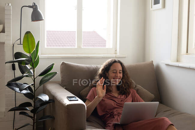 Mujer caucásica pasando tiempo en casa, vistiendo un vestido rosa, sentada en un sofá y sosteniendo su computadora portátil, interactuando usando las redes sociales. Distanciamiento social y autoaislamiento en cuarentena. - foto de stock