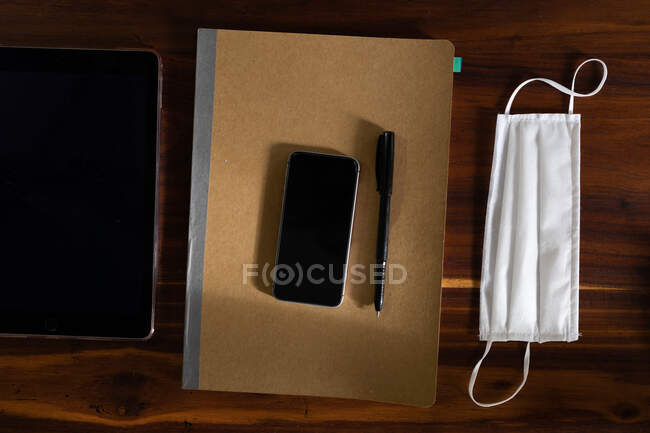 Закройте верхний вид планшета, смартфона, блокнота, ручки и маски на столе во время работы в домашнем офисе. Социальное дистанцирование и самоизоляция в карантинной изоляции. — стоковое фото