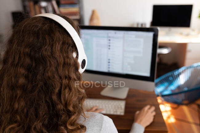 Погляньте на кавказьку жінку, яка проводить вдома, користуючись навушниками, сидить за столом і працює за комп 