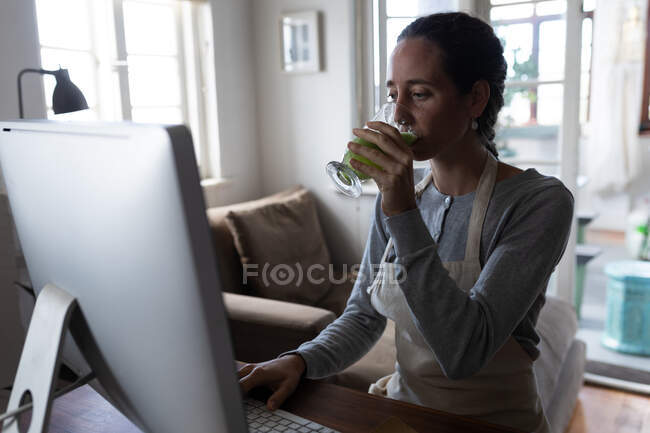 Белая женщина проводит время дома, сидит за столом, работает за компьютером, пьет смузи. Социальное дистанцирование и самоизоляция в карантинной изоляции. — стоковое фото