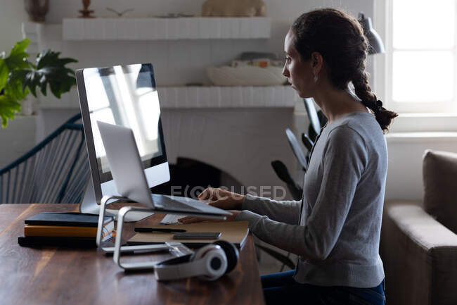 Mujer caucásica pasando tiempo en casa, sentada junto a su escritorio y trabajando usando su computadora. Distanciamiento social y autoaislamiento en cuarentena. - foto de stock