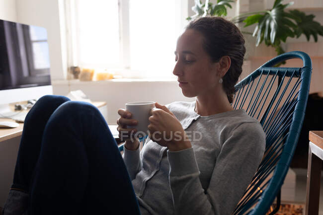 Кавказка проводит время дома, сидит на стуле, держит чашку кофе, расслабляется. Социальное дистанцирование и самоизоляция в карантинной изоляции. — стоковое фото