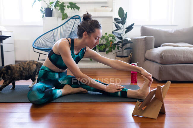 Kaukasische Frau verbringt Zeit zu Hause, trägt Sportbekleidung, sitzt auf einer Yogamatte und streckt sich, nimmt an einem Online-Yoga-Kurs teil, benutzt ihr Tablet. Soziale Distanzierung und Selbstisolierung in Quarantäne. — Stockfoto
