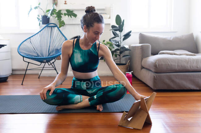 Kaukasische Frau verbringt Zeit zu Hause, trägt Sportbekleidung, sitzt auf einer Yogamatte und streckt sich, nimmt an einem Online-Yoga-Kurs teil, benutzt ihr Tablet. Soziale Distanzierung und Selbstisolierung in Quarantäne. — Stockfoto