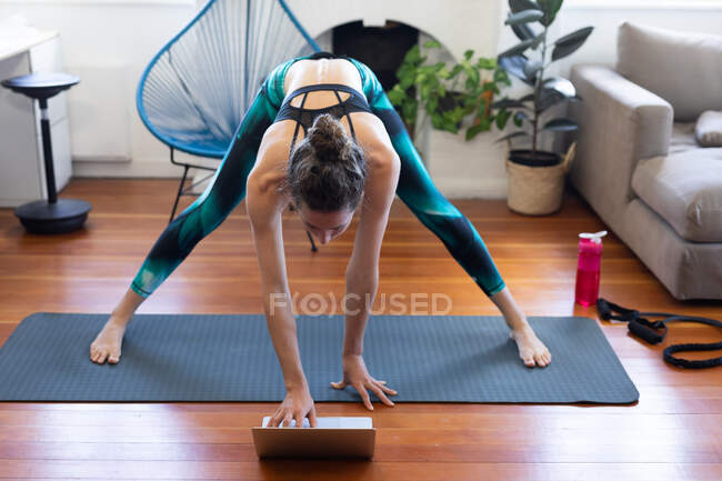 Белая женщина проводит время дома, носит спортивную одежду, тренируется на коврике, присоединяется к онлайн-курсу йоги, использует свой ноутбук. Социальное дистанцирование и самоизоляция в карантинной изоляции. — стоковое фото