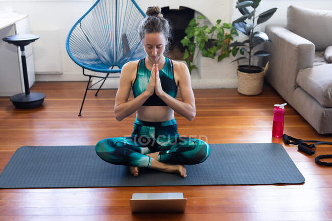 Белая женщина проводит время дома, одетая в спортивную одежду, сидит на коврике и медитирует, присоединяется к онлайн-курсу йоги, пользуется ноутбуком. Социальное дистанцирование и самоизоляция в карантинной изоляции. — стоковое фото
