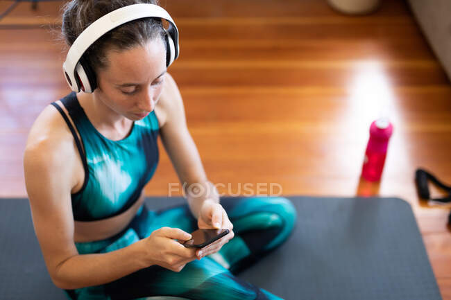 Caucasian woman spending time at home, wearing sportswear and headphones, sitting on a mat and using her smartphone. Social distancing and self isolation in quarantine lockdown. — Stock Photo